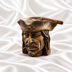 Antique bronze pirate head ashtray with dark distressed detailing and a skull-and-crossbones hat, holding a cigar on white satin fabric.