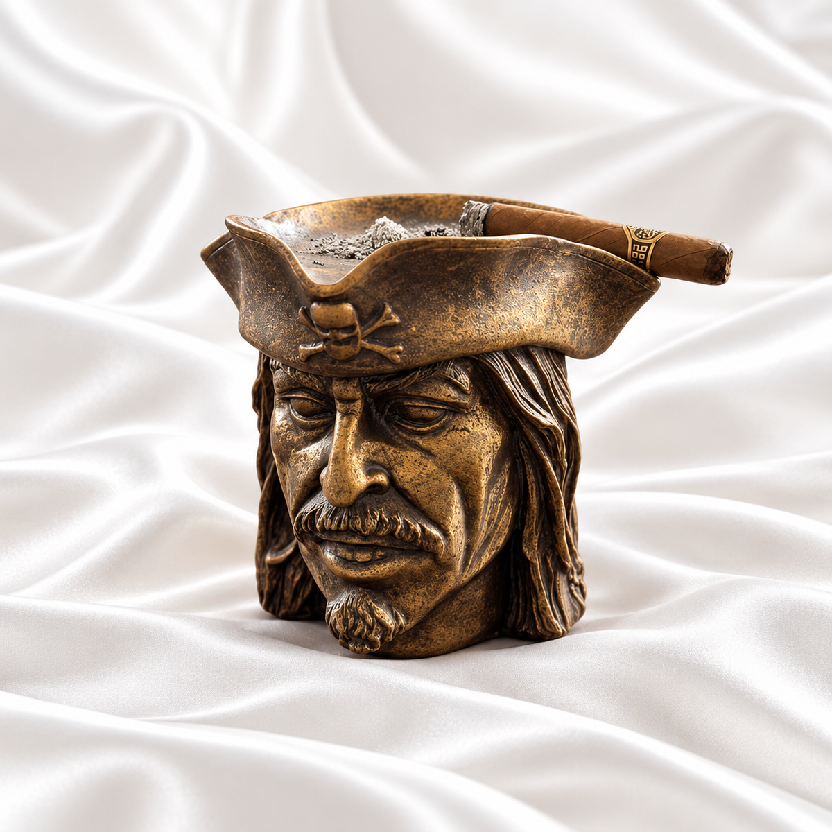 Antique bronze pirate head ashtray with dark distressed detailing and a skull-and-crossbones hat, holding a cigar on white satin fabric.