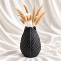9.5-inch matte black sculpted leaf vase with beige dried stems on a white satin fabric background.