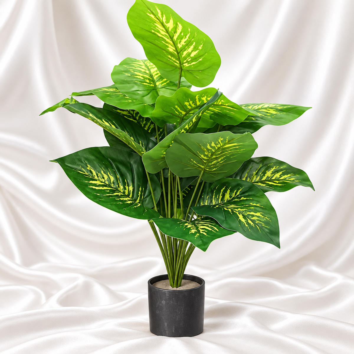 Artificial variegated green plant with broad leaves in a black cylindrical pot against a white satin fabric background.