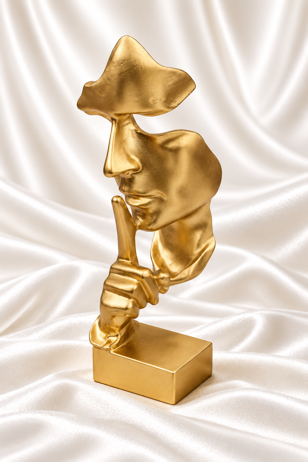 Matte gold abstract shushing face sculpture with square base on white satin fabric.