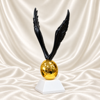 Black angel wings sculpture with detailed feathers, a reflective gold-tone egg and white pedestal, displayed against off-white satin fabric.