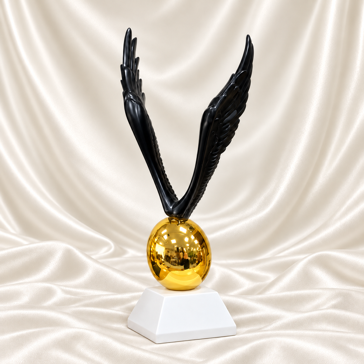 Black angel wings sculpture with detailed feathers, a reflective gold-tone egg and white pedestal, displayed against off-white satin fabric.