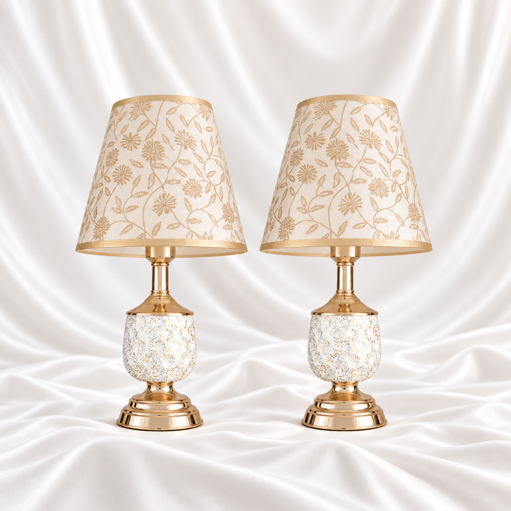 Pair of white and gold-speckled table lamps with gold-tone bases and cream floral shades, displayed against a white satin fabric background.
