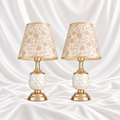 Pair of white and gold-speckled table lamps with gold-tone bases and cream floral shades, displayed against a white satin fabric background.