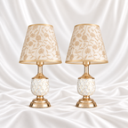 Pair of white and gold-speckled table lamps with gold-tone bases and cream floral shades, displayed against a white satin fabric background.