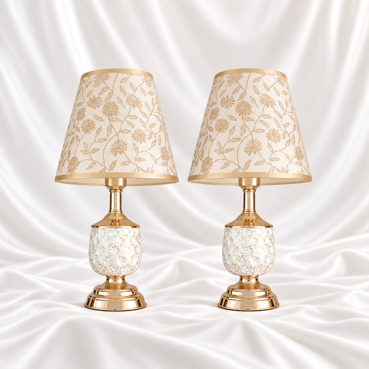 Pair of white and gold-speckled table lamps with gold-tone bases and cream floral shades, displayed against a white satin fabric background.