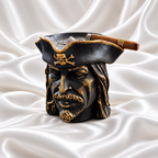 Black pirate head ashtray with gold facial accents, gold hat trim and a skull-and-crossbones emblem, holding a cigar on white satin fabric.