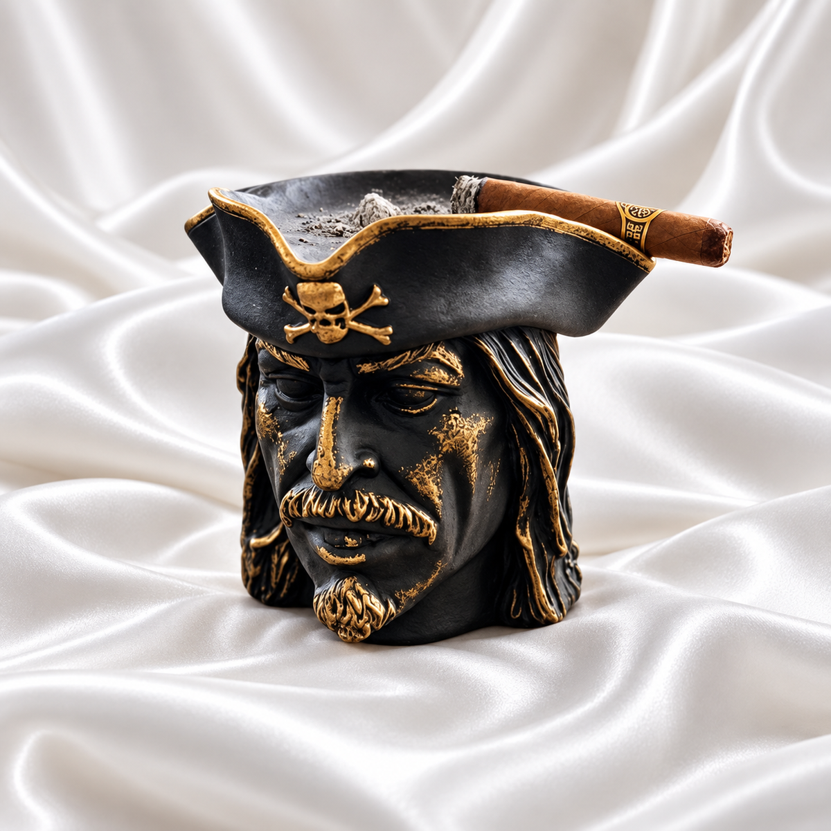 Black pirate head ashtray with gold facial accents, gold hat trim and a skull-and-crossbones emblem, holding a cigar on white satin fabric.