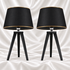 Pair of black tripod table lamps with black tapered shades and gold-tone trim, displayed against a white satin fabric background.