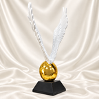White angel wings sculpture with detailed feathers, a reflective gold-tone egg and black pedestal, displayed against off-white satin fabric.