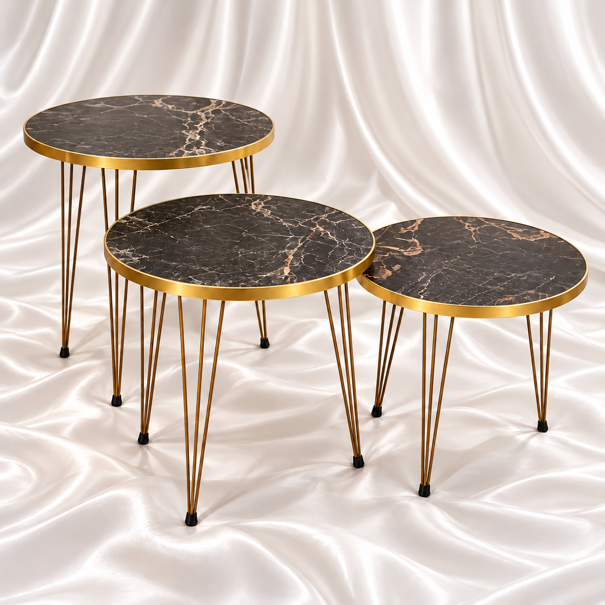 Set of three round black marble-effect side tables with gold-tone rims and slender hairpin legs on a white satin fabric background.