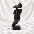 Black abstract face sculpture making a finger-to-lips gesture on a rectangular base against white satin fabric.