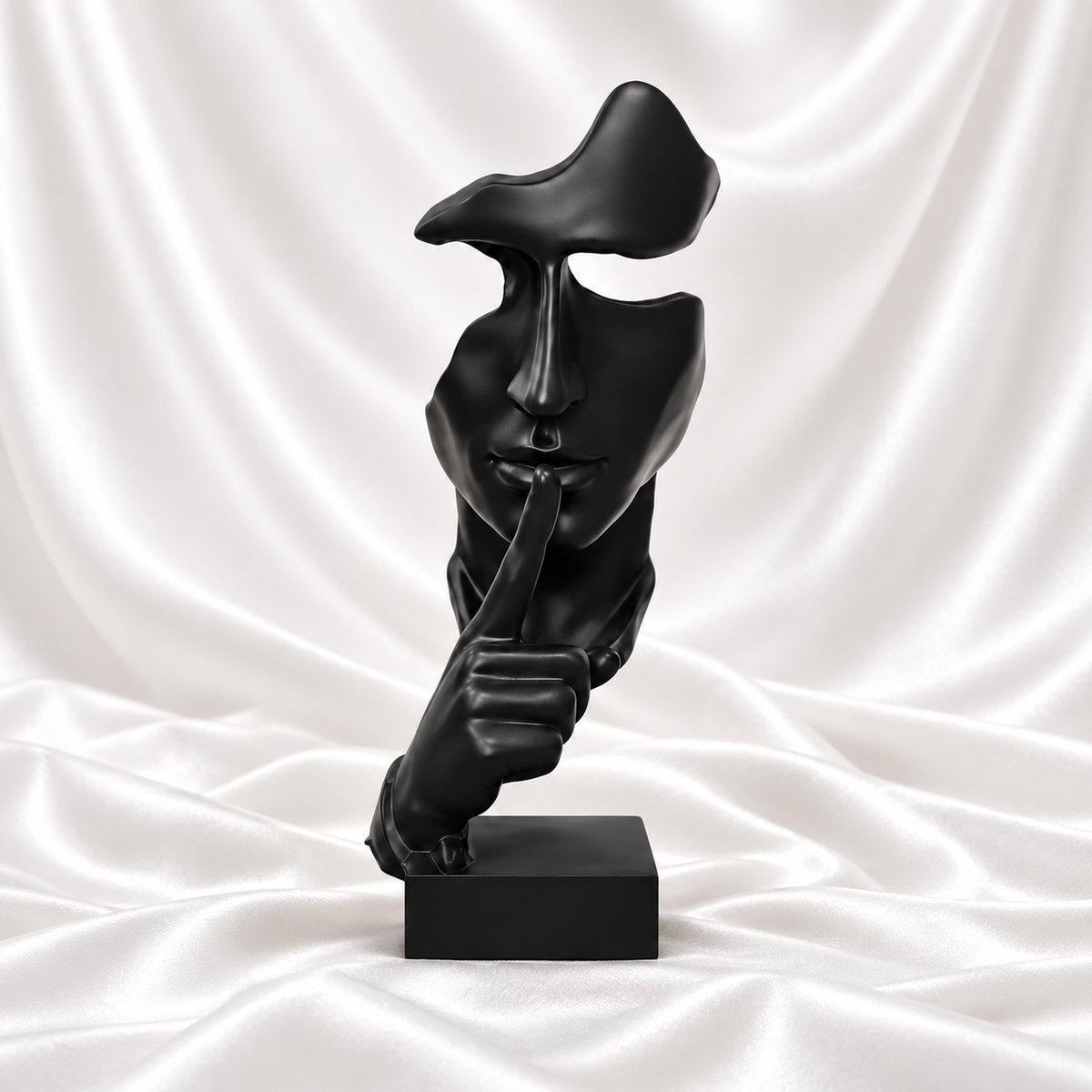 Black abstract face sculpture making a finger-to-lips gesture on a rectangular base against white satin fabric.