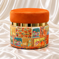 Single orange upholstered ottoman stool with a gold-tone band and colourful patchwork base on a white satin background.