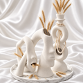 Set of three white textured vases featuring two abstract faces and one double-ring design, styled with dried wheat and a pearl necklace on a marble-pattern tray against white satin.