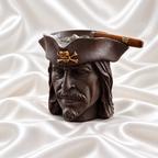 Dark brown pirate head ashtray with sculpted facial details and a gold skull-and-crossbones emblem, holding a cigar on white satin fabric.