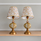 Pair of gold-tone mosaic table lamps with white and beige leaf-patterned shades, clear decorative collars and round bases on a wooden shelf.