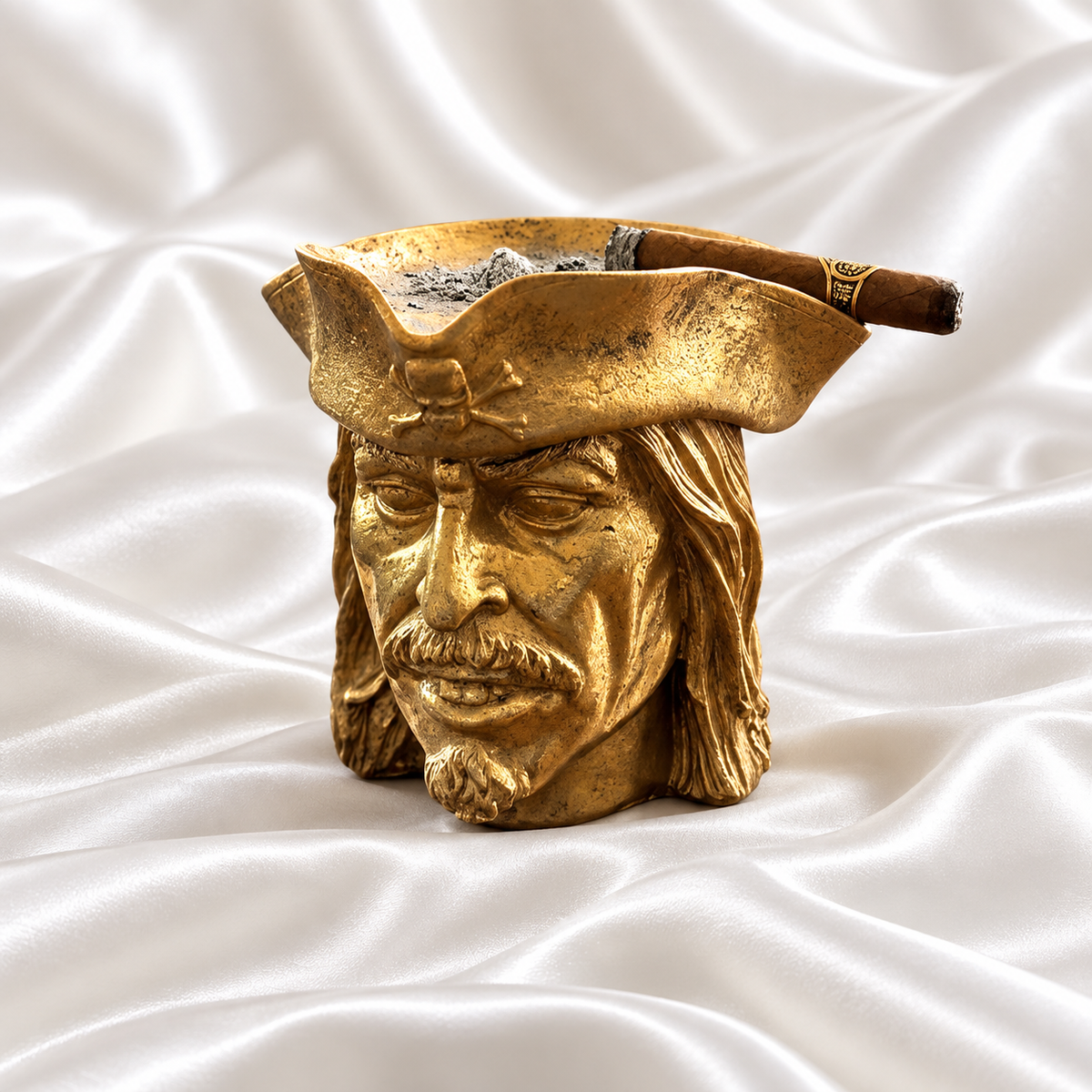 Gold pirate head ashtray with detailed facial features and a skull-and-crossbones hat, holding a cigar on white satin fabric.