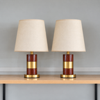 Pair of brown woodgrain-look table lamps with gold-tone centre bands, round gold bases and cream tapered shades on a wooden shelf against a light grey wall.