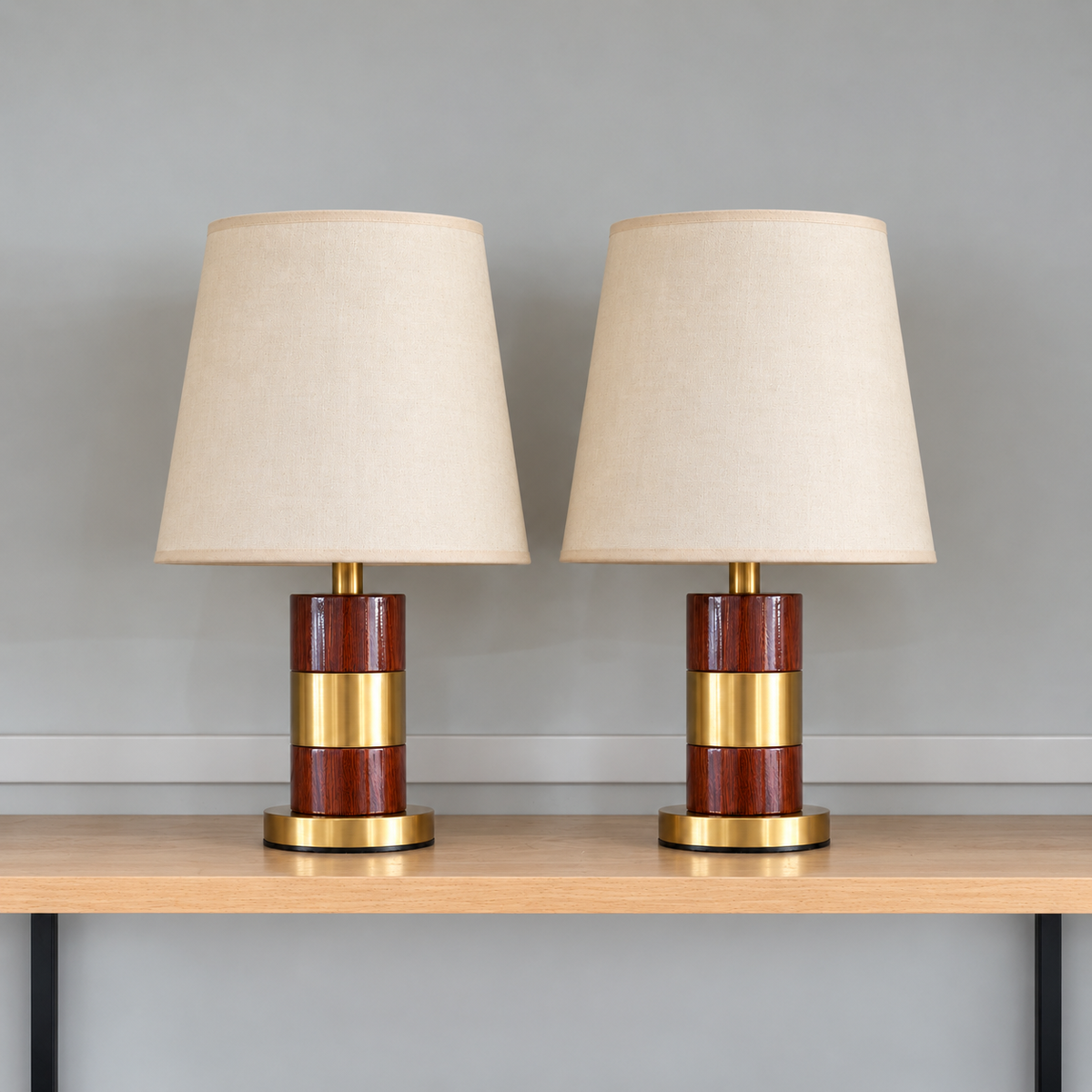 Pair of brown woodgrain-look table lamps with gold-tone centre bands, round gold bases and cream tapered shades on a wooden shelf against a light grey wall.