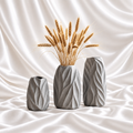 Set of three grey decorative vases with sculpted leaf patterns, displayed with dried wheat against a white satin fabric background.