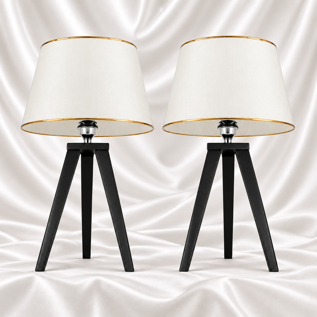 Pair of black tripod table lamps with white tapered shades and gold-tone trim on a white satin fabric background.