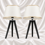 Pair of black tripod table lamps with white tapered shades and gold-tone trim on a white satin fabric background.