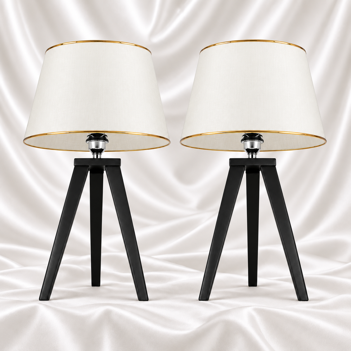 Pair of black tripod table lamps with white tapered shades and gold-tone trim on a white satin fabric background.