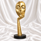 Gold abstract face and hand sculpture with a textured gold stem and round black base, displayed against white satin fabric.