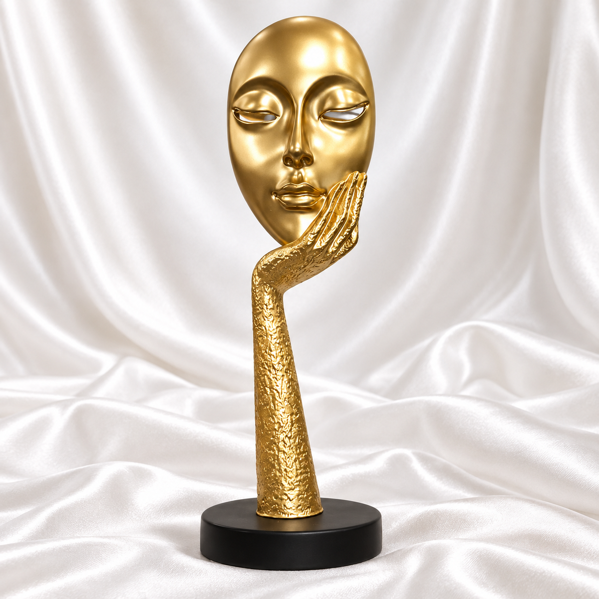 Gold abstract face and hand sculpture with a textured gold stem and round black base, displayed against white satin fabric.