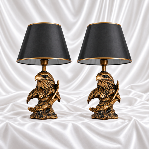 Pair of matching antique gold and black eagle table lamps with tapered black shades and gold trim, displayed on white satin fabric.
