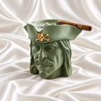 Sage green pirate head ashtray with a gold skull-and-crossbones emblem on its hat, holding a cigar on white satin fabric.