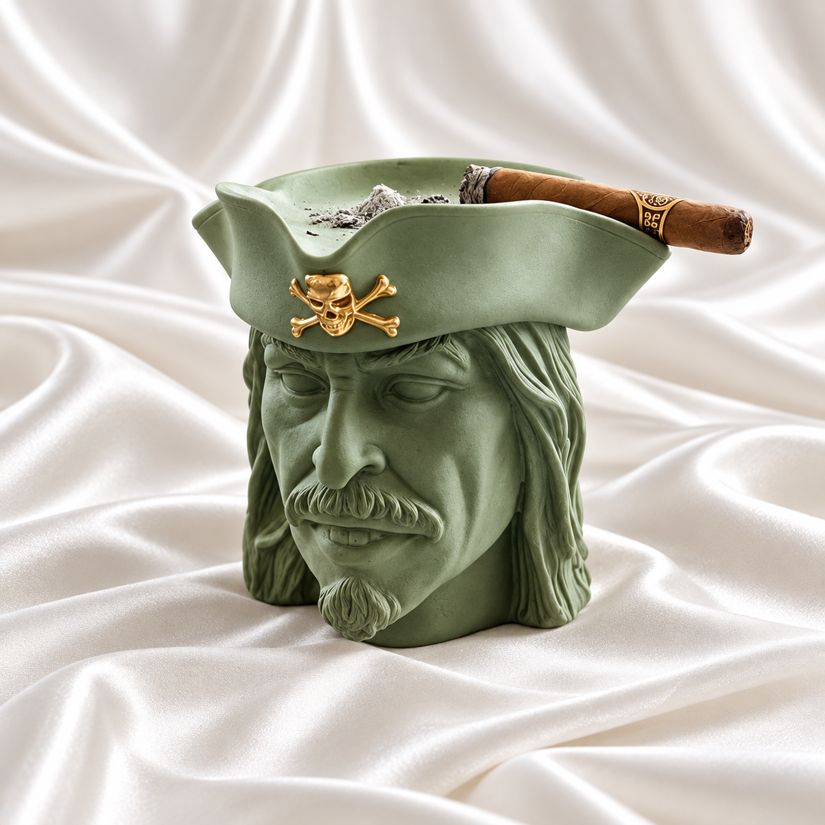 Sage green pirate head ashtray with a gold skull-and-crossbones emblem on its hat, holding a cigar on white satin fabric.