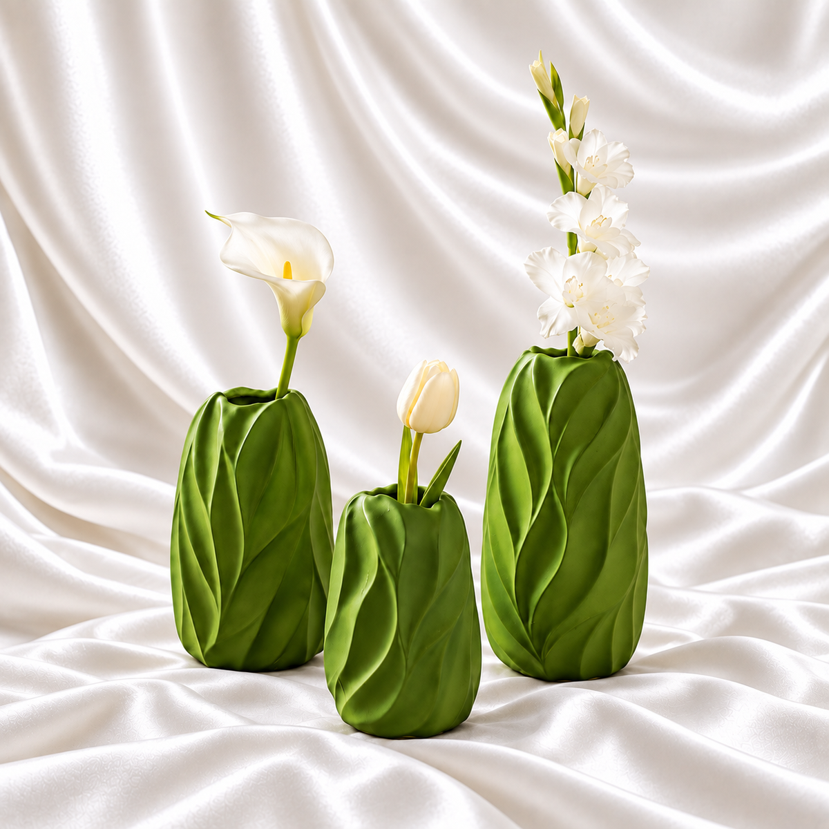Set of three leaf green decorative vases in graduated sizes with raised leaf patterns and white flowers, displayed on white satin fabric.
