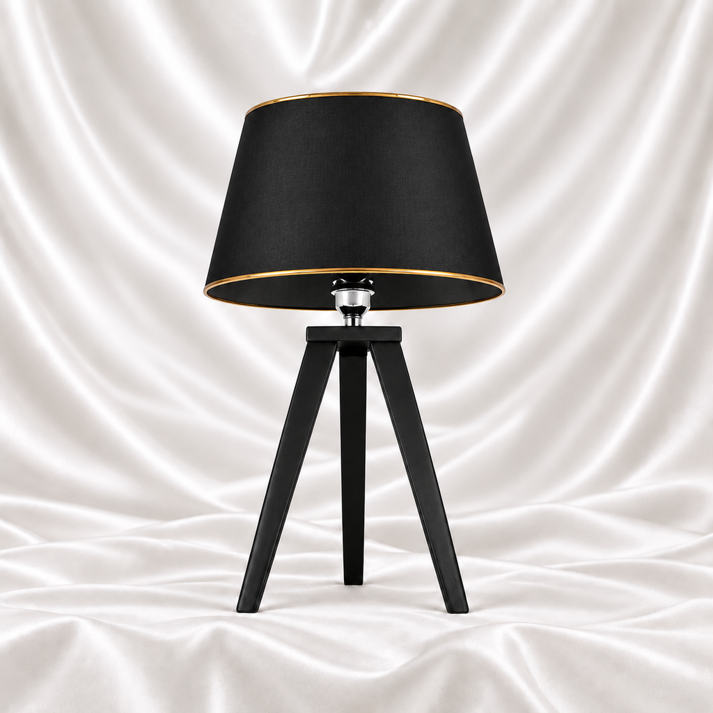Black tripod table lamp with a black tapered shade and gold-tone trim on a white satin fabric background.