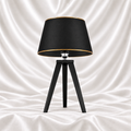 Black tripod table lamp with a black tapered shade and gold-tone trim on a white satin fabric background.