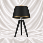 Black tripod table lamp with a black tapered shade and gold-tone trim on a white satin fabric background.
