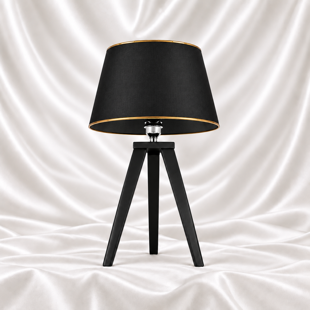 Black tripod table lamp with a black tapered shade and gold-tone trim on a white satin fabric background.