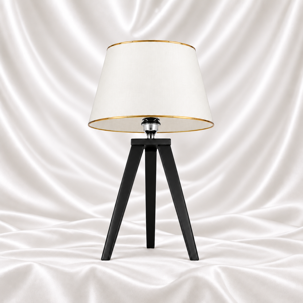 Black tripod table lamp with a white tapered shade and gold-tone trim on a white satin fabric background.