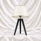 Black tripod table lamp with a white tapered shade and gold-tone trim on a white satin fabric background.